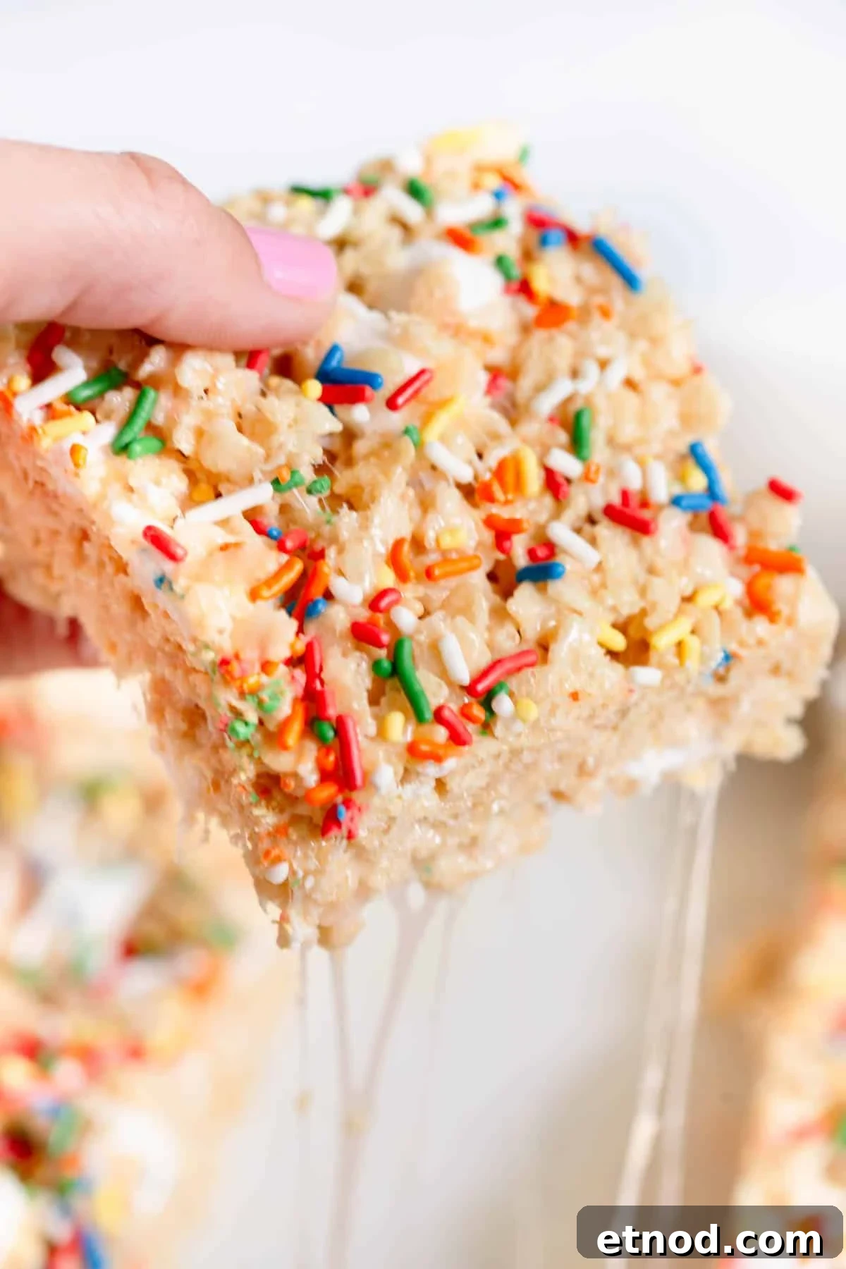 A hand with pink fingernails hold a Birthday Cake Rice Krispie Treat that has gooey marshmallow sticking to the other piece.