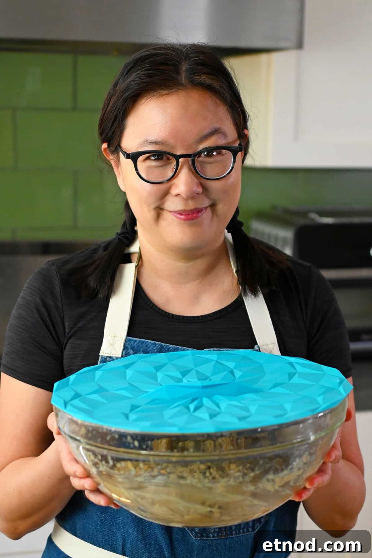 Soft Banana Chocolate Chip Cookies — Paleo Grain-Free Recipe 11 A smiling Asian woman in glasses is holding a glass mixing bowl filled with paleo banana cookie batter covered with a blue silicone lid.