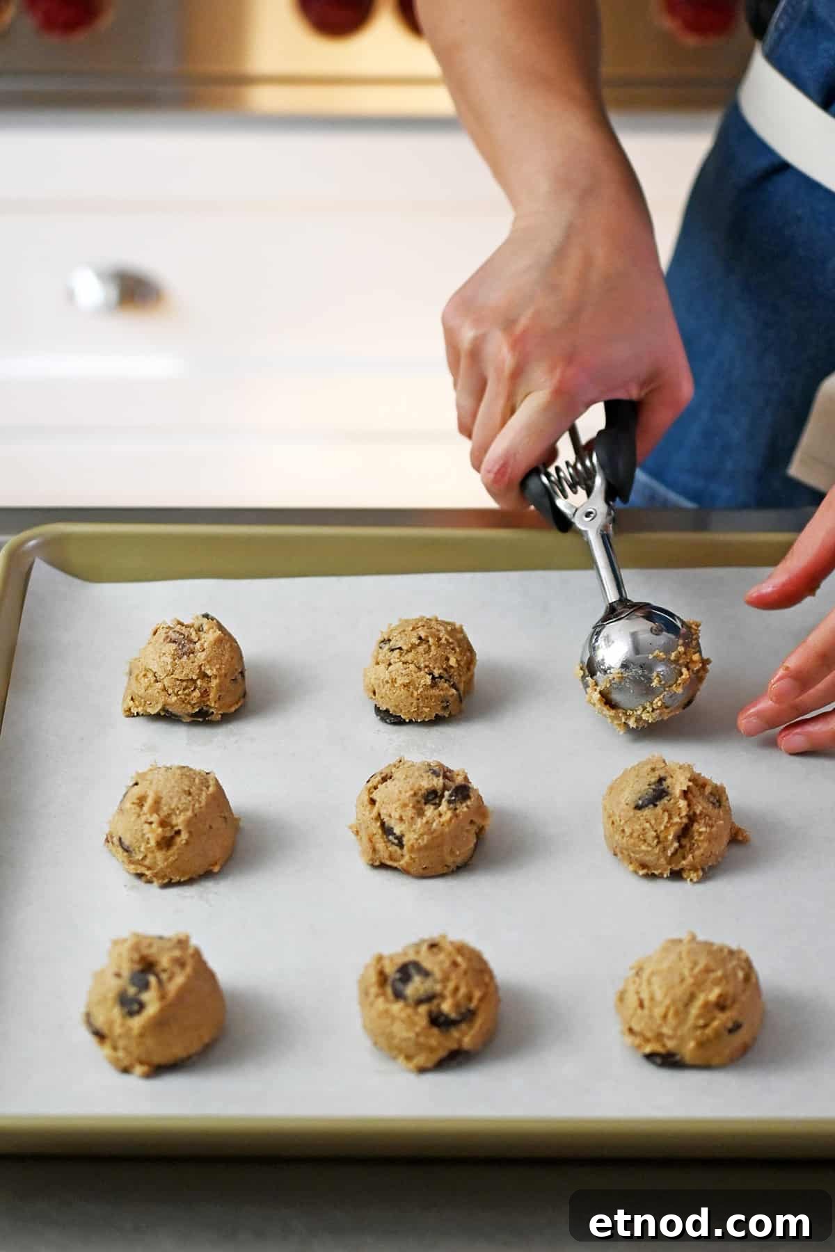 Soft Banana Chocolate Chip Cookies — Paleo Grain-Free Recipe 12 A rimmed baking sheet lined with parchment paper is covered with raw banana cookie dough balls being portioned out with a medium ice cream scooper.