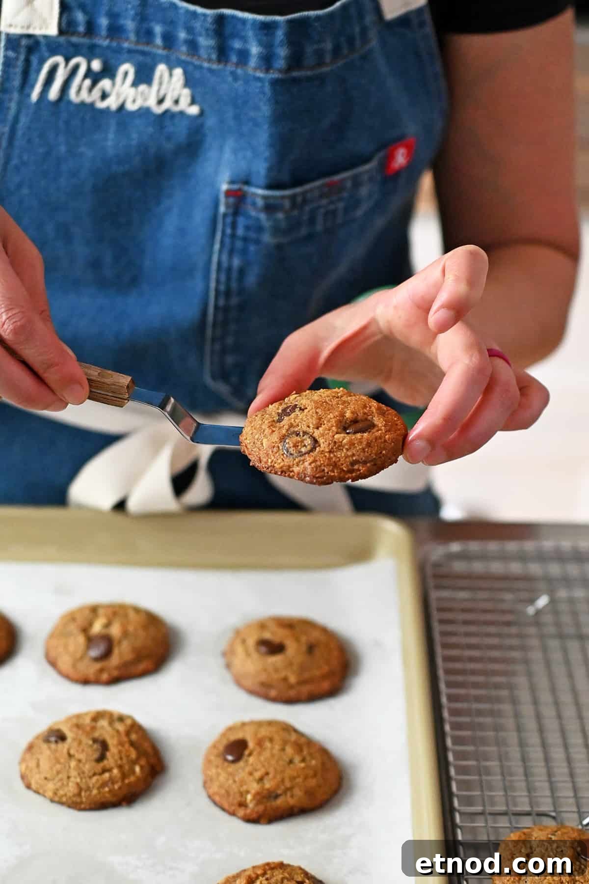 Soft Banana Chocolate Chip Cookies — Paleo Grain-Free Recipe 15 A person in a blue apron is using a small offset spatula to transfer freshly baked paleo banana cookies from a baking pan to a wire cooling rack,