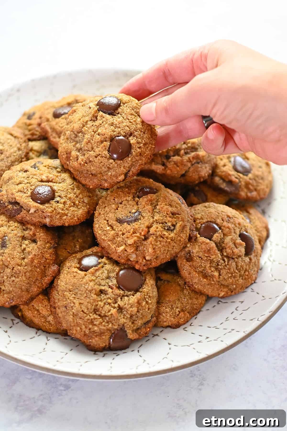 Soft Banana Chocolate Chip Cookies — Paleo Grain-Free Recipe 17 A hand is grabbing a paleo banana chocolate chip cookie from a white plate piled hight with cookies.