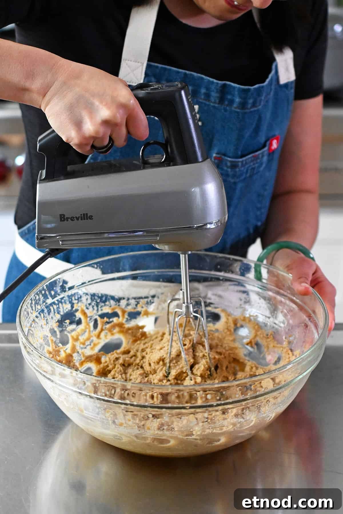 Soft Banana Chocolate Chip Cookies — Paleo Grain-Free Recipe 9 A person in a blue apron is using a hand mixer to combine the batter for paleo banana cookies in a glass mixing bowl.