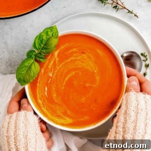 Creamy Roasted Red Pepper Soup with Garlic and Basil 18 two hands grabbing a white bowl full of creamy roasted red pepper soup, garnished with basil, heavy cream, and thyme