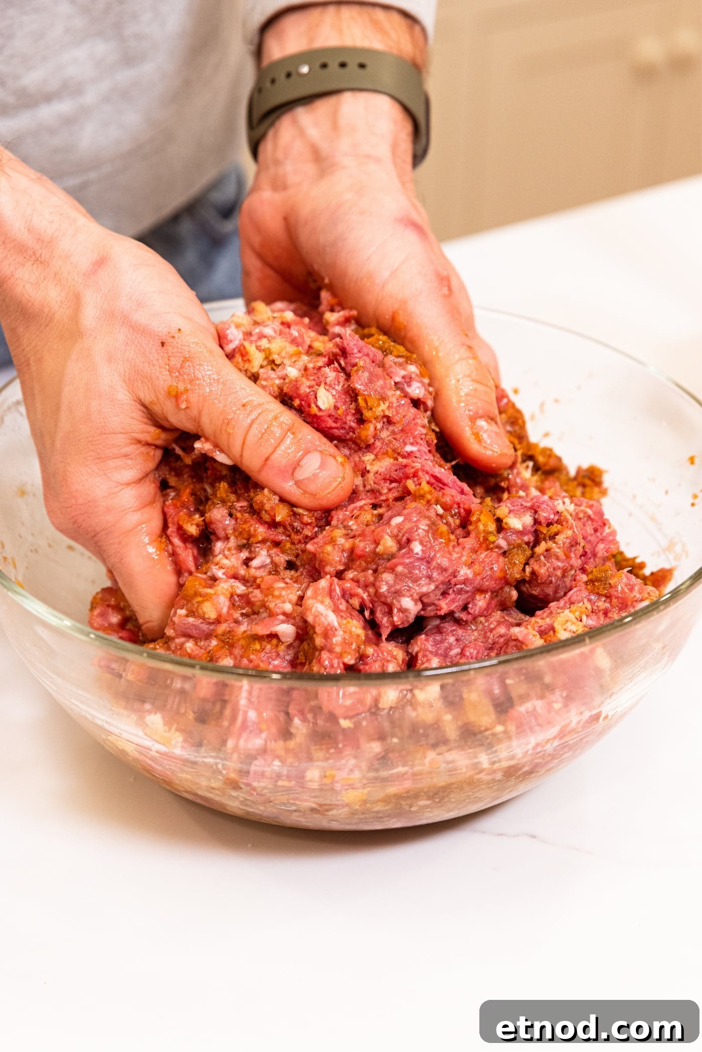 Classic Comfort Meatloaf Recipe for Perfect Weeknight Dinners 13 Hands pushing together ingredients needed for meatloaf including beef, pork, and breadcrumbs in glass bowl on white countertop.