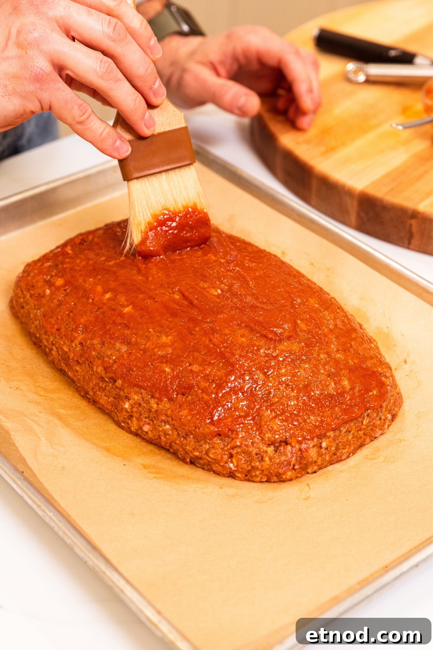 Classic Comfort Meatloaf Recipe for Perfect Weeknight Dinners 16 Hand using brush to apply ketchup mixture on the top of meatloaf sitting on parchment-lined baking sheet.
