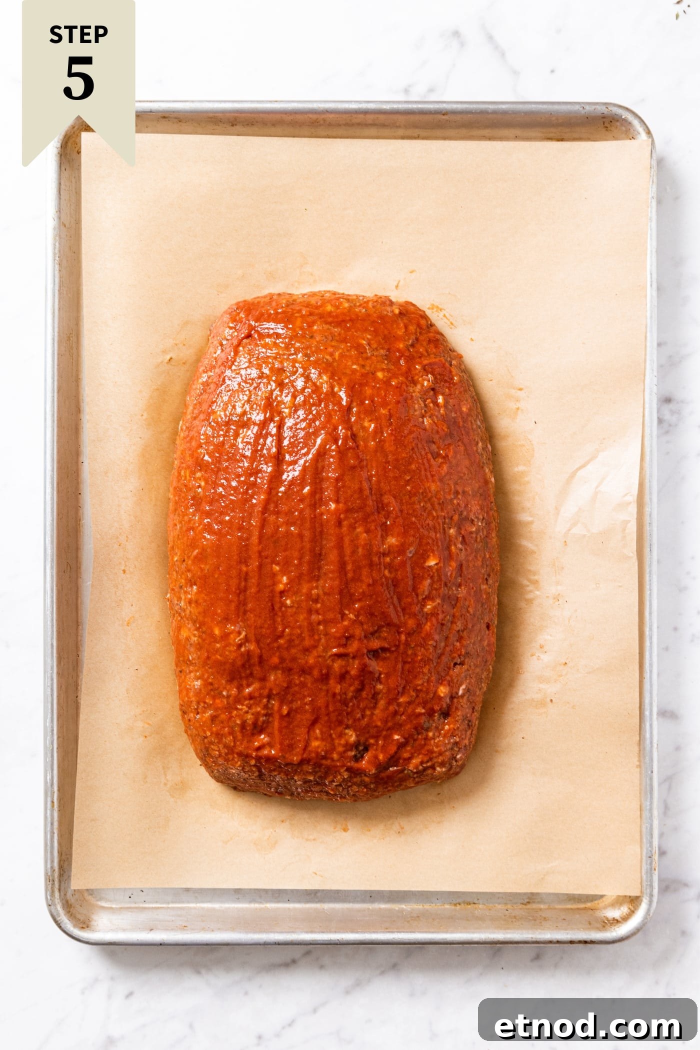 Classic Comfort Meatloaf Recipe for Perfect Weeknight Dinners 17 Parchment-lined baking sheet with unbaked meatloaf with ketchup brushed over the top sitting on marble surface.
