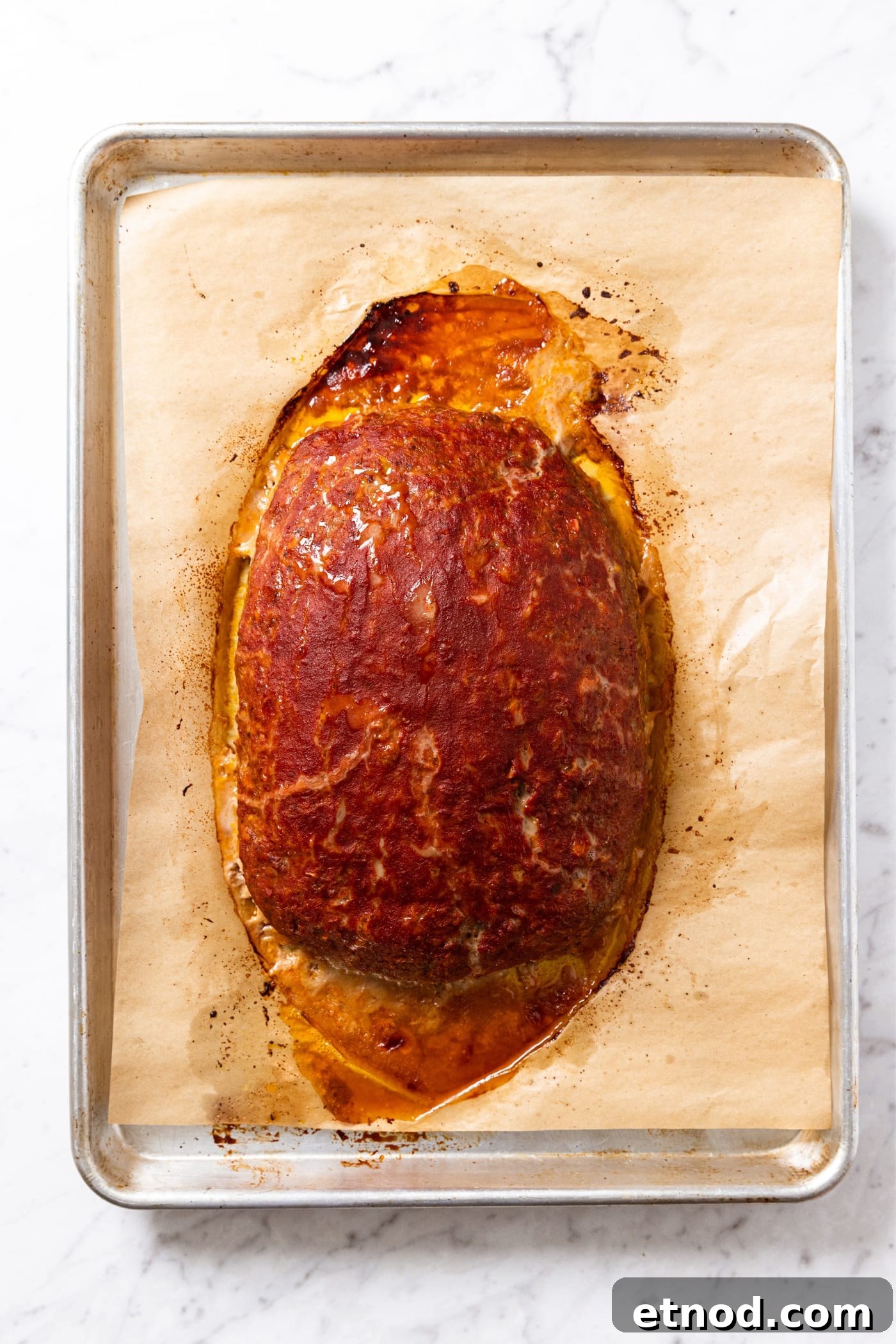 Classic Comfort Meatloaf Recipe for Perfect Weeknight Dinners 18 Baked meatloaf sitting on parchment-lined baking sheet with browned ketchup mixture and excess grease from meat seeping out.