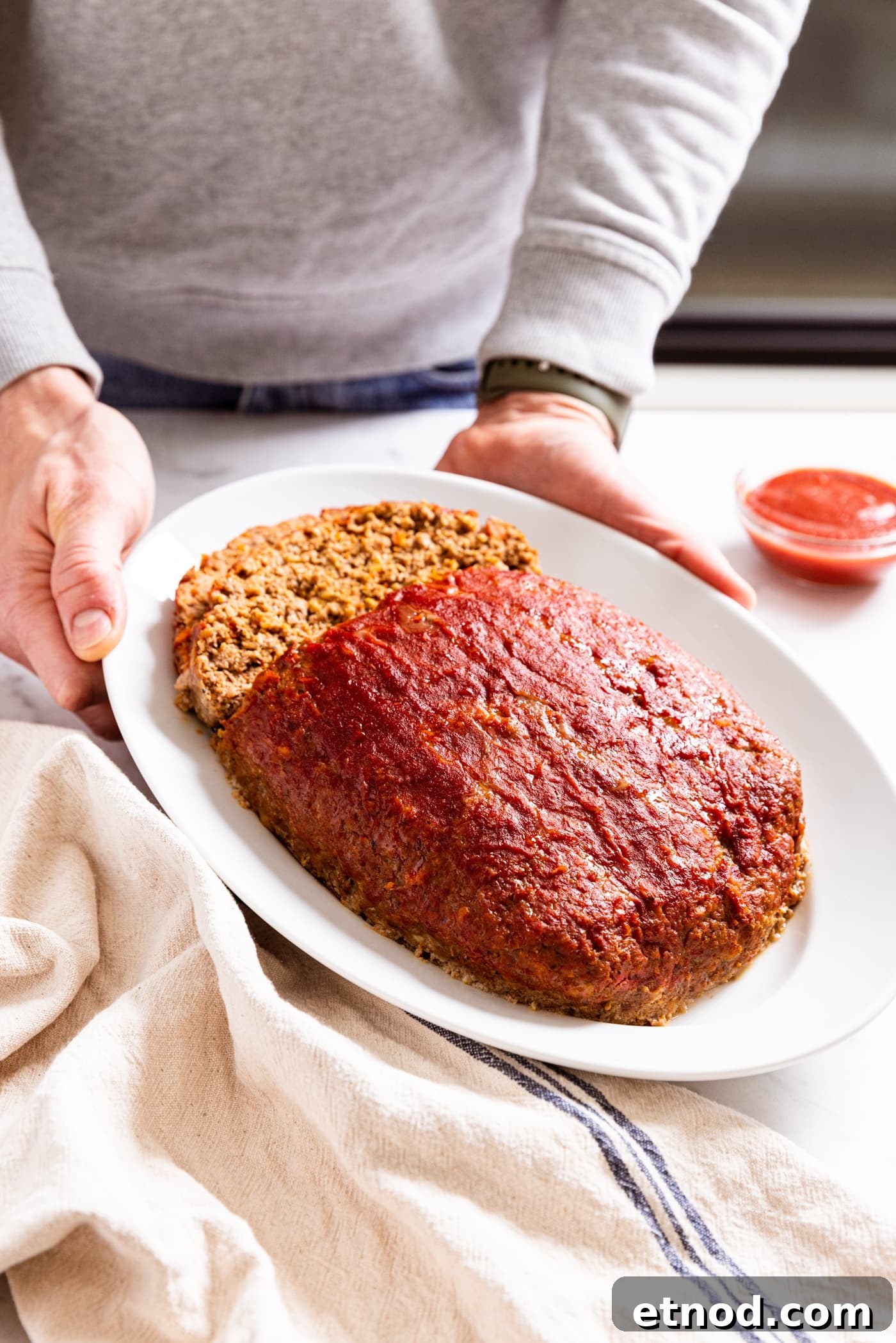 Classic Comfort Meatloaf Recipe for Perfect Weeknight Dinners 19 Hands holding white platter with meatloaf with two slices sliced off the back side with small bowl filled with ketchup to the side.