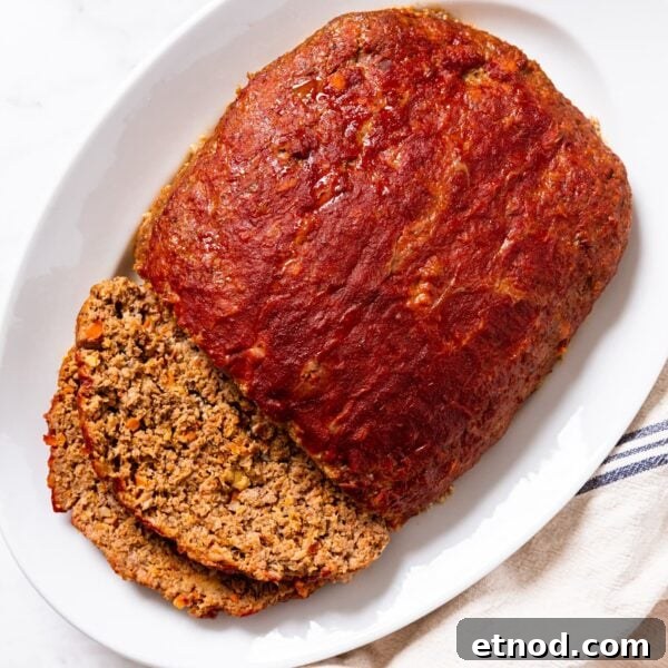 Classic Comfort Meatloaf Recipe for Perfect Weeknight Dinners 20 White platter sitting on white marble surface filled with meatloaf with two slices sliced off the side with ketchup all over the top.