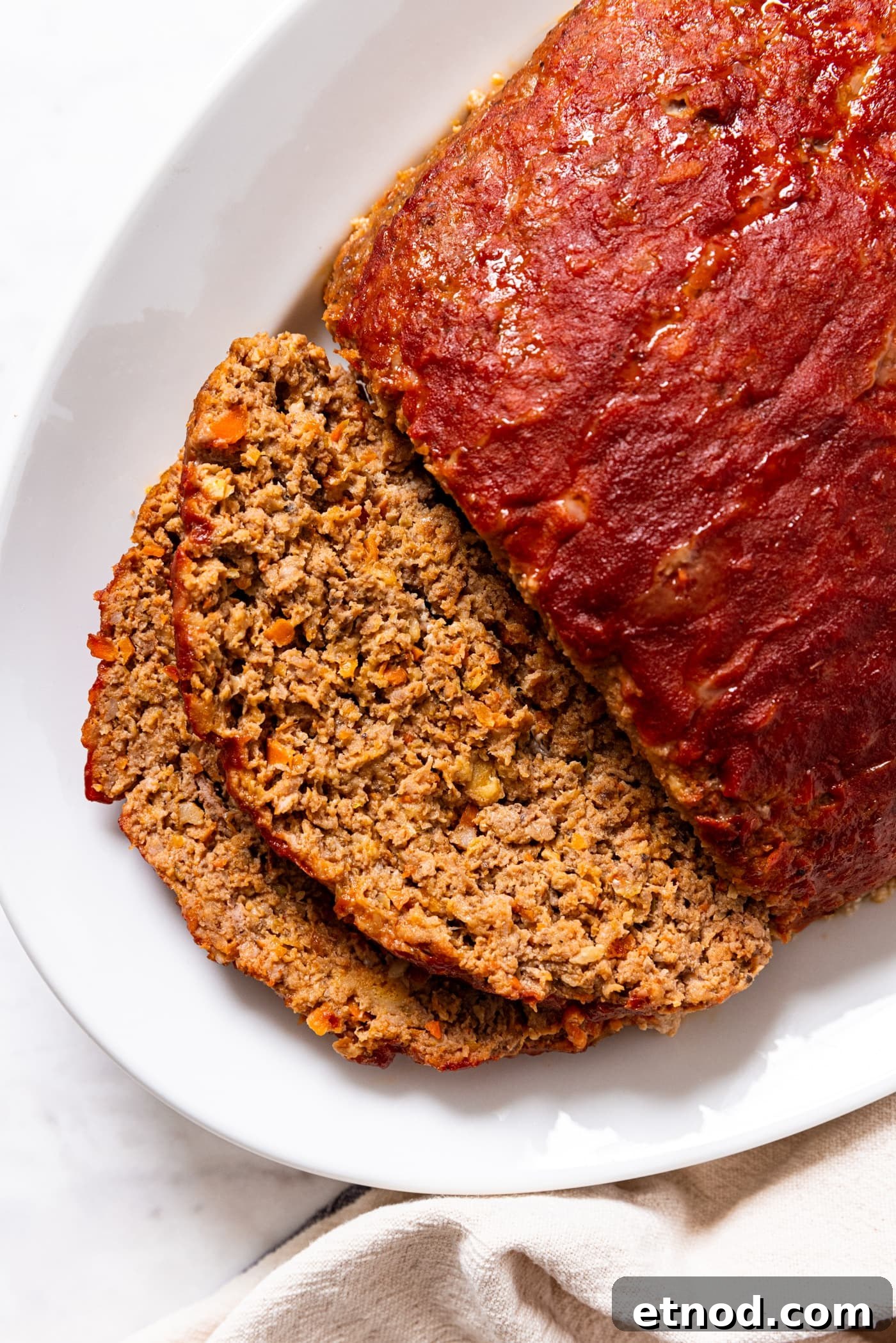 Classic Comfort Meatloaf Recipe for Perfect Weeknight Dinners 3 White serving platter with meatloaf sitting with two slices sliced off the front with ketchup slathered all over the top showing interior texture of meat.