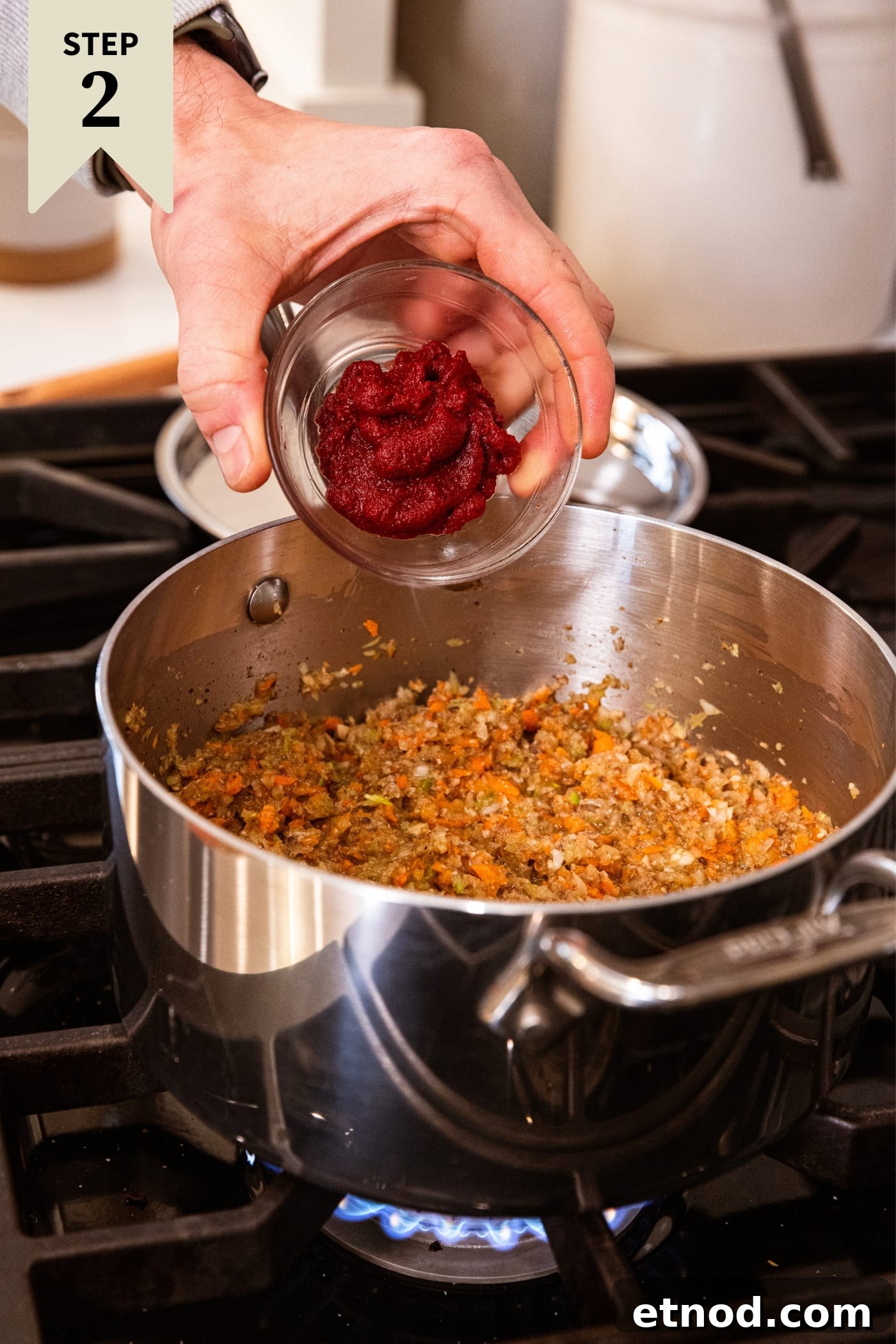 Classic Comfort Meatloaf Recipe for Perfect Weeknight Dinners 9 Hand holding small glass bowl about to pour tomato paste into vegetable mixture in a metal stockpot on the heat on stovetop.