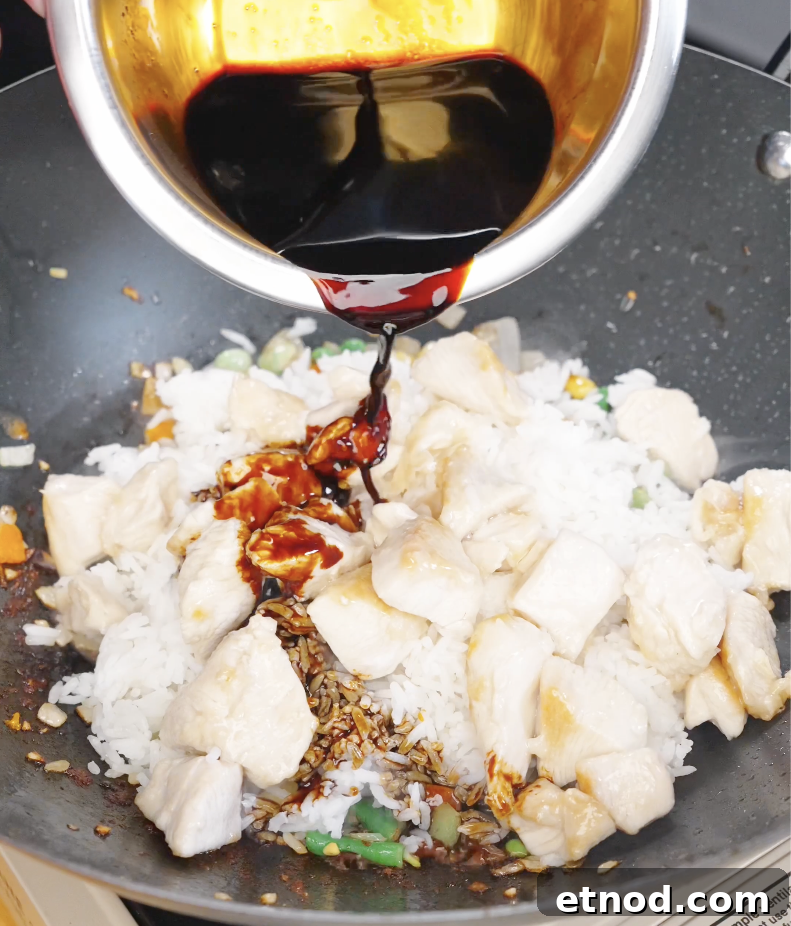 Soy-based sauce being poured over rice and cooked chicken in a pan, coating the fried rice with flavor.