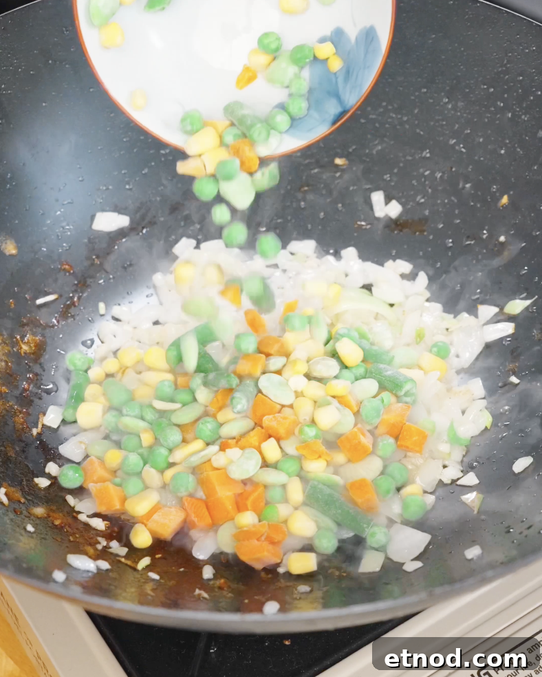 Frozen mixed vegetables being added to sautéed onions in a pan, adding color and texture to chicken fried rice.