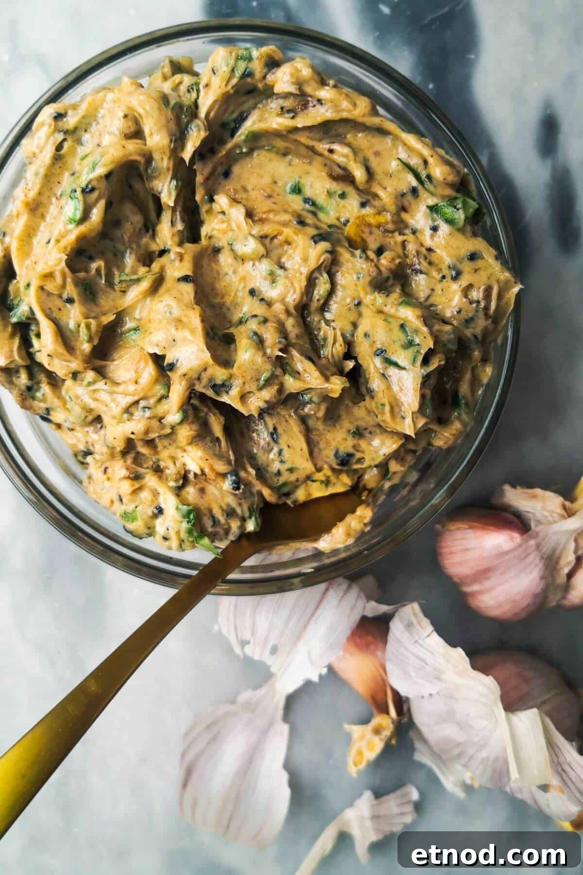 Roasted Garlic Butter Recipe: Creamy Spread for Bread & Cooking 2 Roasted garlic butter in a small glass bowl with a gold spoon inside, with garlic cloves in the background.