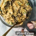 Roasted garlic butter in a small glass bowl with a gold spoon inside, with garlic cloves in the background.