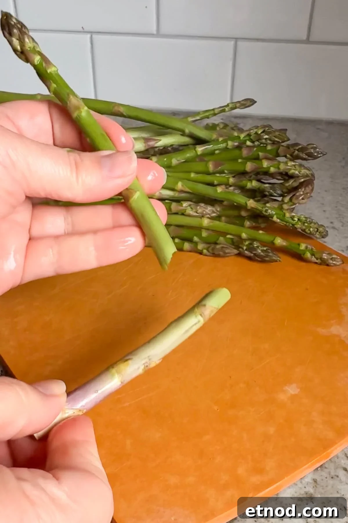 Grilled Asparagus Without Foil: Simple, Crispy Recipe Under 20 Minutes 4 A hand breaks an asparagus stalk near its base over an orange cutting board, with a bunch of asparagus in the background on a kitchen counter.