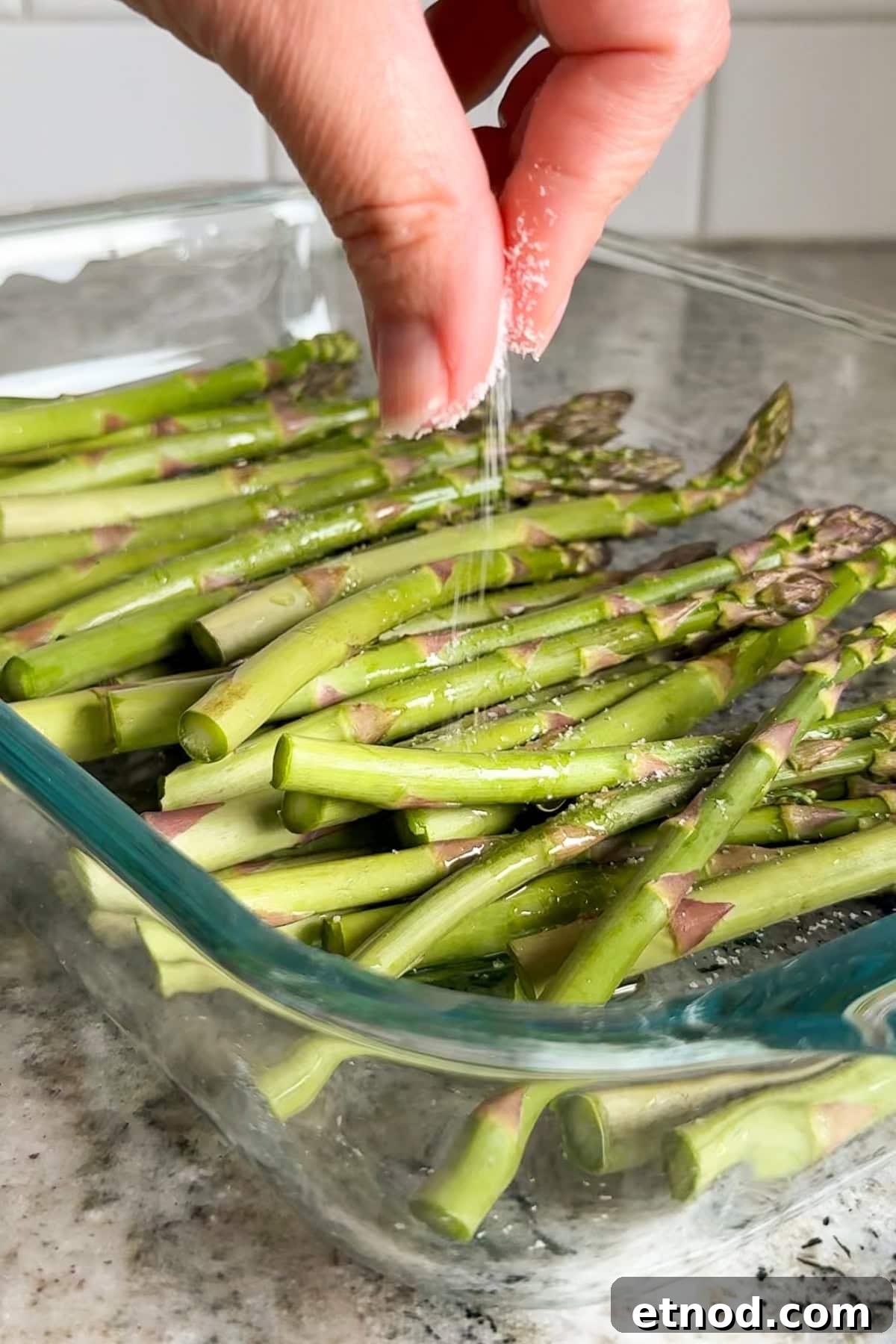Grilled Asparagus Without Foil: Simple, Crispy Recipe Under 20 Minutes 6 A hand sprinkles salt over fresh asparagus spears arranged in a clear glass baking dish on a kitchen countertop.
