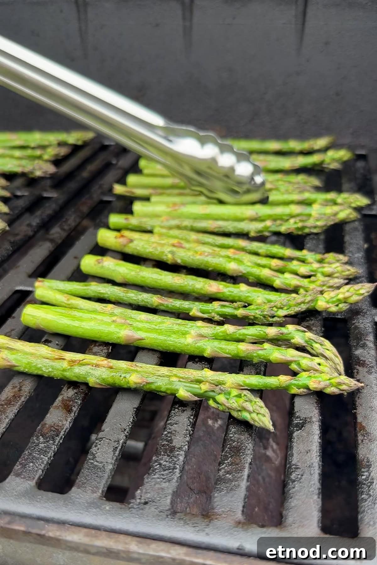 Grilled Asparagus Without Foil: Simple, Crispy Recipe Under 20 Minutes 8 A close-up of asparagus spears being grilled on an outdoor barbecue. A pair of metal tongs is about to pick up one of the asparagus spears. The grill grates are visibly heated.