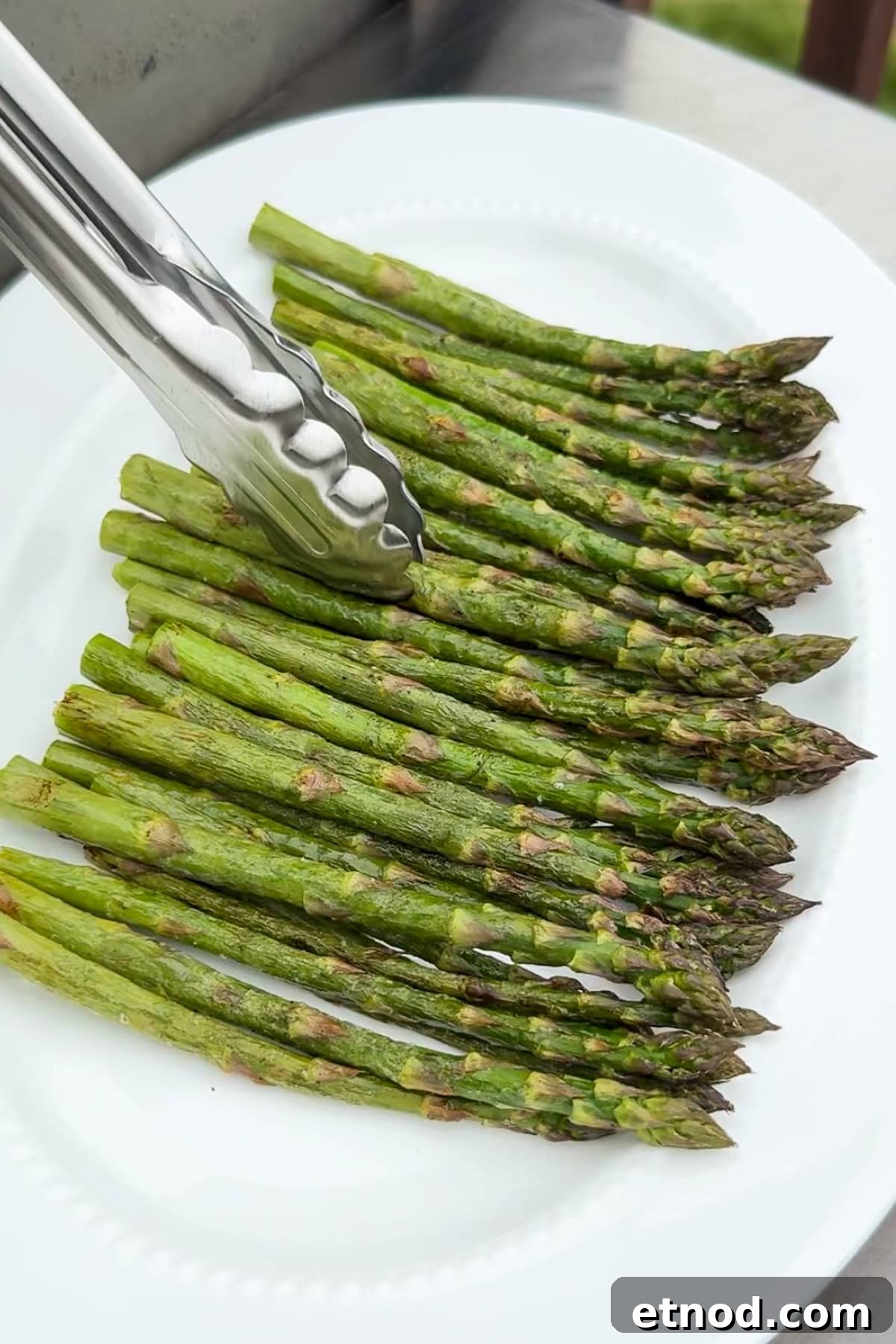 Grilled Asparagus Without Foil: Simple, Crispy Recipe Under 20 Minutes 9 A pair of metal tongs is picking up grilled asparagus spears arranged in a neat row on a white plate.