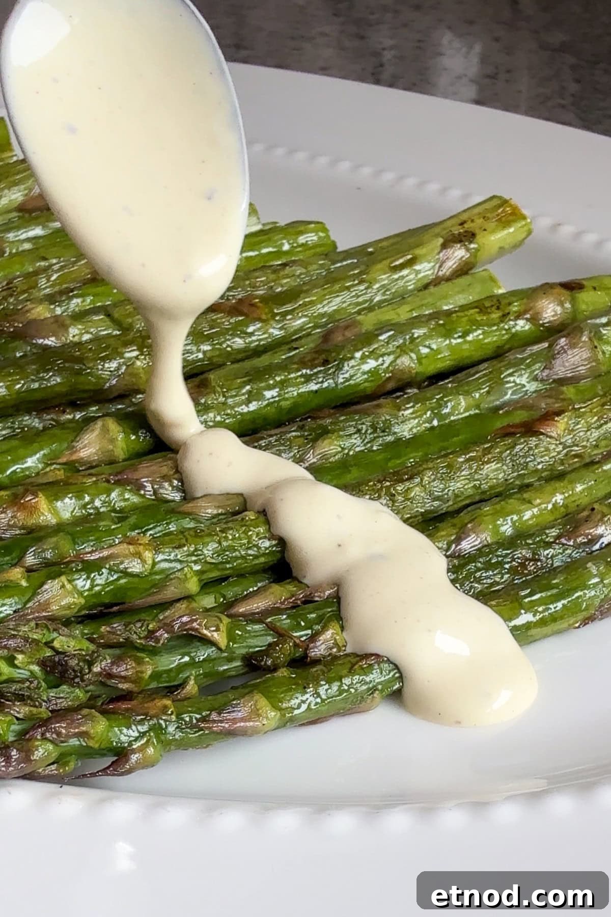Grilled Asparagus Without Foil: Simple, Crispy Recipe Under 20 Minutes 10 A spoon pours a creamy, light-colored sauce over a serving of roasted asparagus spears arranged on a white plate.