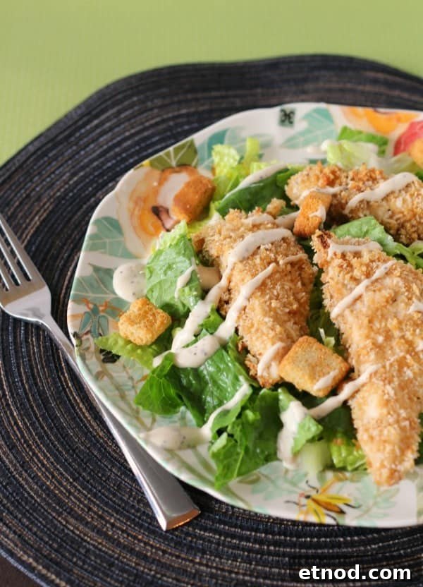 Crispy Caesar Chicken Tenders with Creamy Parmesan Dressing 2 Partial image of chicken tenders served on a salad with dressing drizzled over all, on decorative plate with fork.