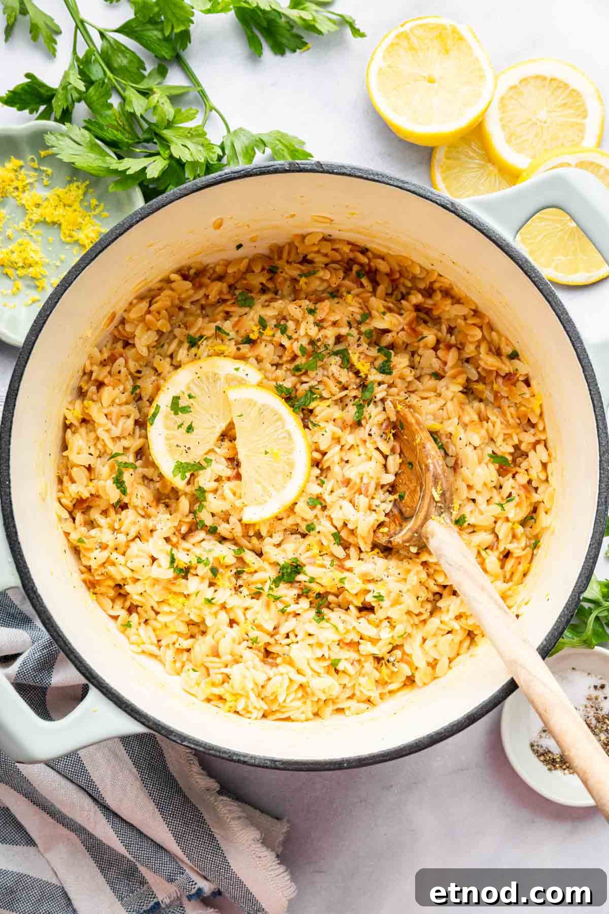 Creamy Lemon Orzo with Parmesan and Fresh Herbs 8 Lemon orzo in a white pan with a wooden spoon and lemon slices on the side.