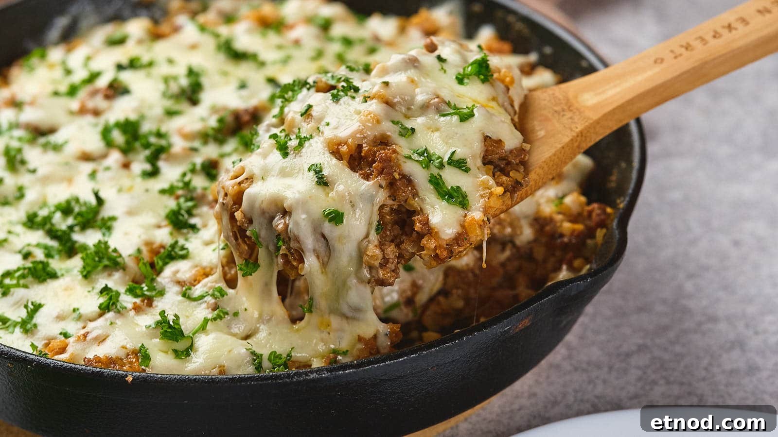 A wooden spoon lifting a serving of italian ground beef skillet garnished with parsley from a cast-iron skillet.