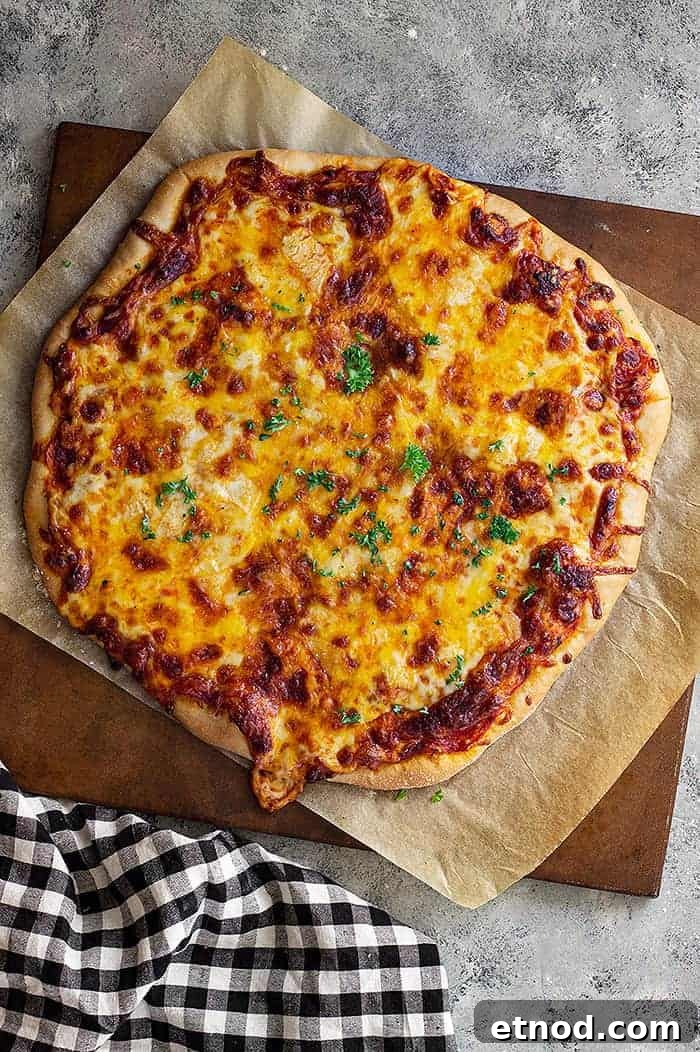 overhead: homemade cheese pizza on parchment paper on a wooden cutting board with a black and white checkered towel to the left