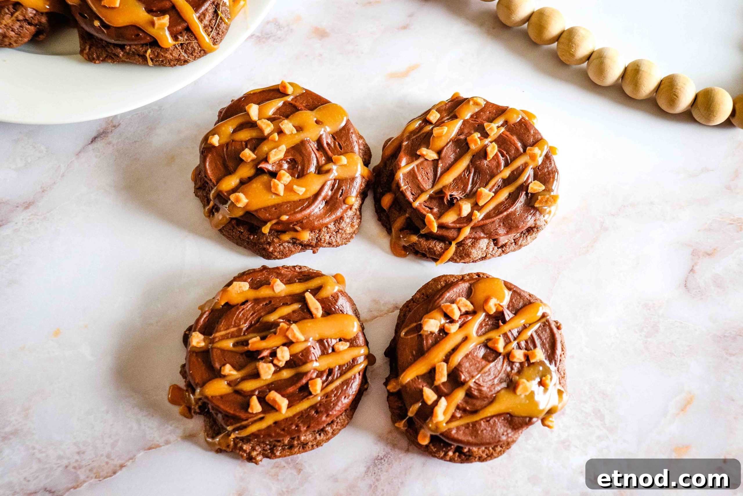 21 Quirky-Looking Desserts That Disappear First at Parties 2 Chocolate Caramel Cookies with frosting, caramel drizzle, and chopped nuts on a white marble surface.