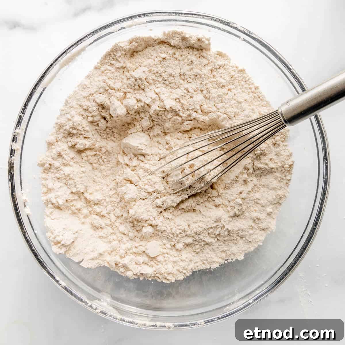 Pumpkin Banana Bread Recipe with Warm Spices and Moist Crumb 4 A glass bowl filled with flour mixture and a whisk on a white surface.