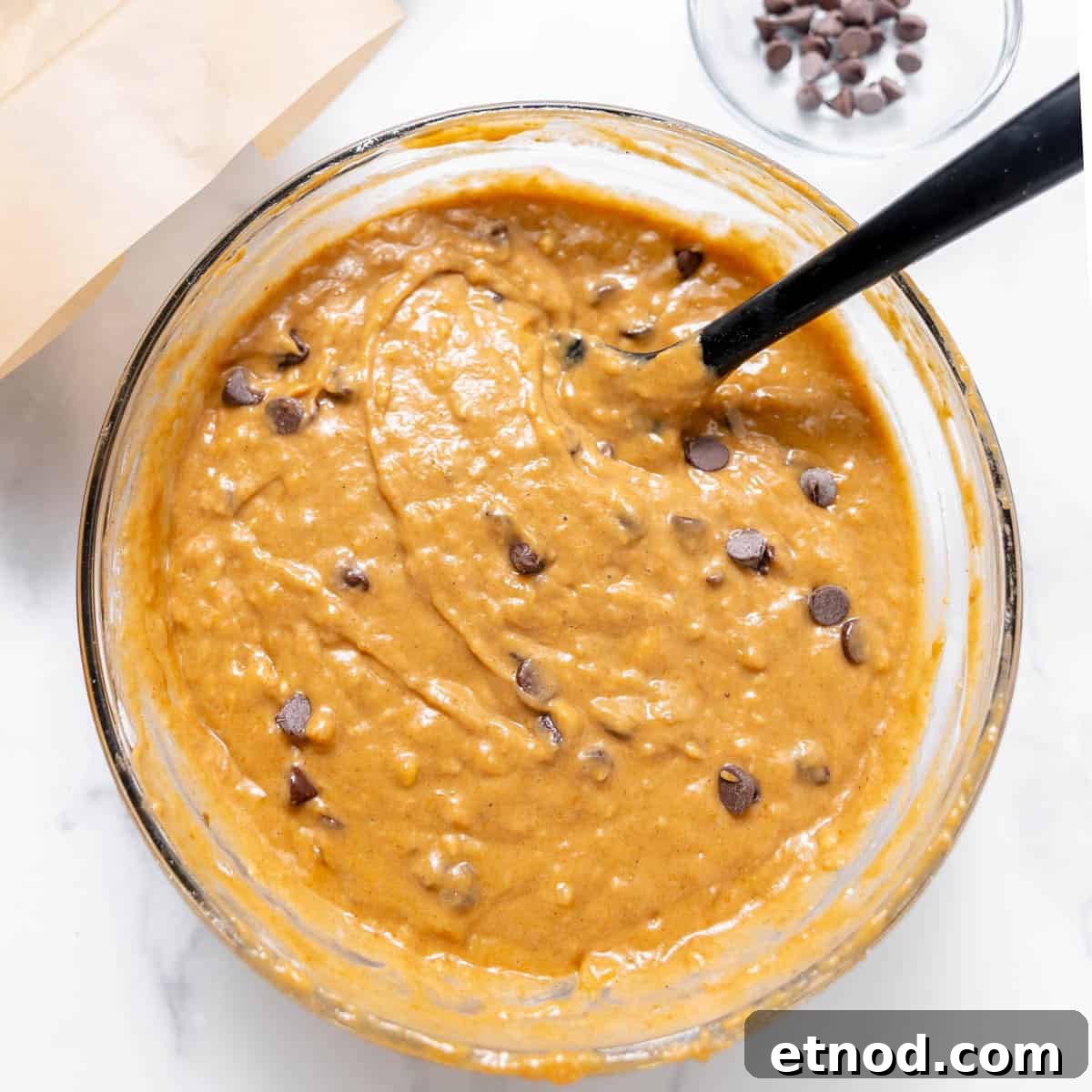 Pumpkin Banana Bread Recipe with Warm Spices and Moist Crumb 6 A glass bowl filled with pumpkin bread batter mixed with chocolate chips. A black spatula is in the bowl, and a small bowl of chocolate chips is nearby.