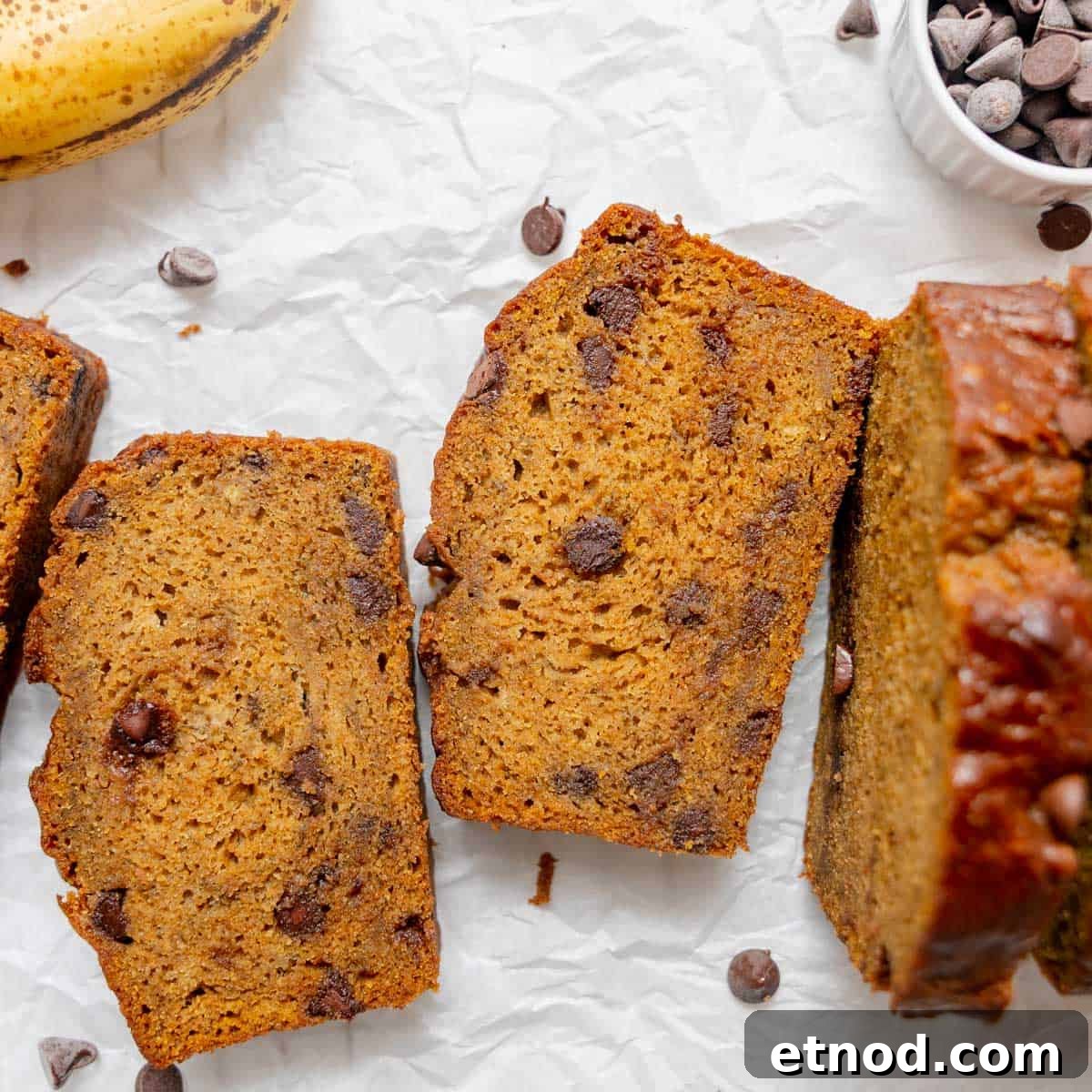 Pumpkin Banana Bread Recipe with Warm Spices and Moist Crumb 10 Sliced chocolate chip banana bread on parchment paper, with a banana and a bowl of chocolate chips nearby.