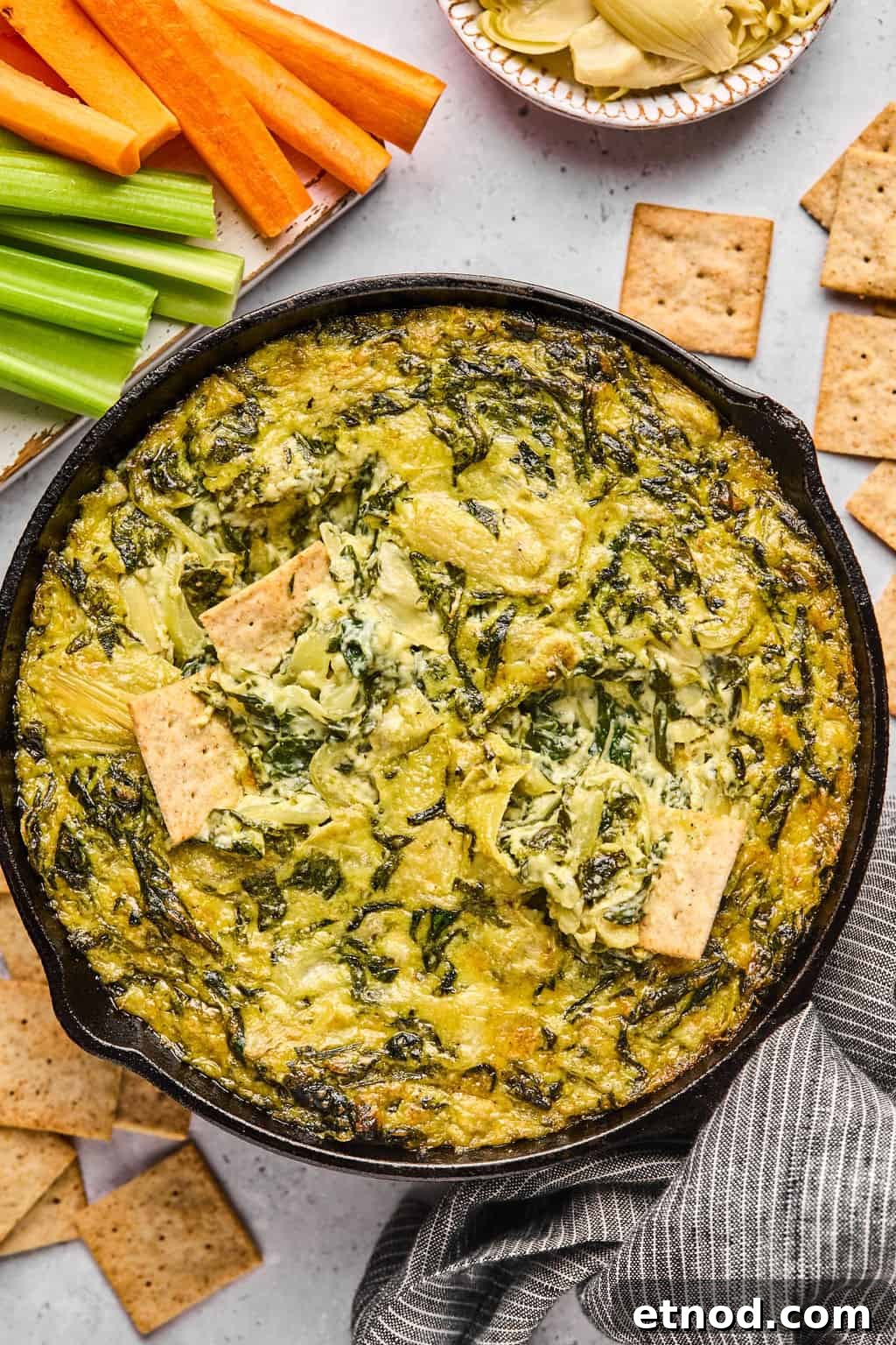 dairy free spinach artichoke dip in a skillet with crackers and veggies on the side.